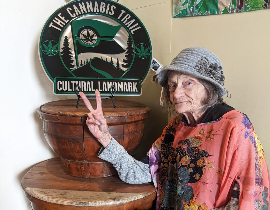 Pebbles Trippet gives a peace sign in front of the Cannabis Trail Cultural Landmark sign that honors her advocacy work at Sol de Mendocino.